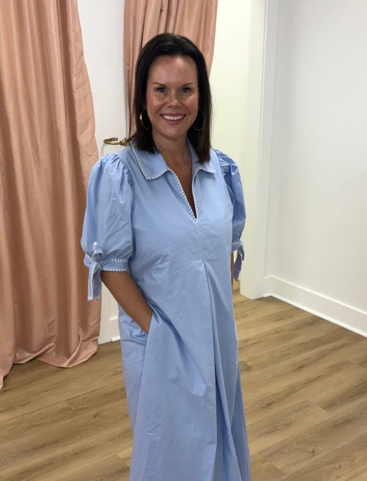Woman wearing a light blue dress standing in a room with pink curtains and wooden flooring.